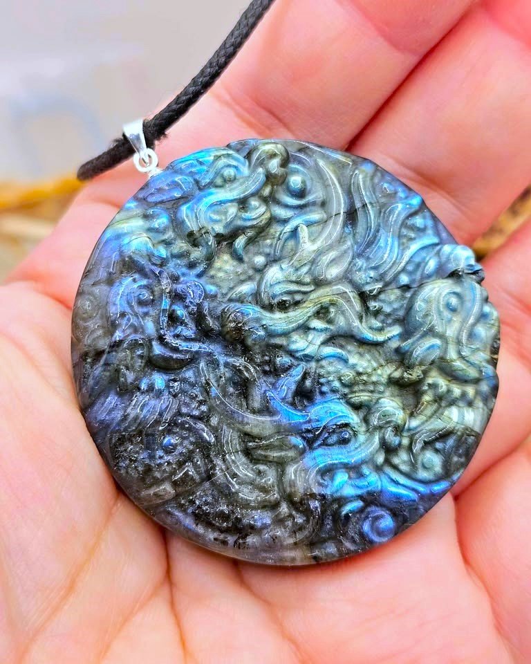 A round carved labradorite pendant with a detailed dragon design, held in a person's hand. The pendant features natural blue and green iridescent flashes on a dark gray base, hanging from a black cord with a silver bail.