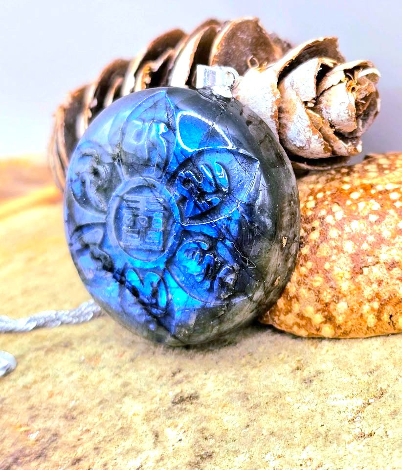 A round blue labradorite pendant with carved Celtic-style symbols, resting on a textured stone surface next to a pinecone and a silver chain.