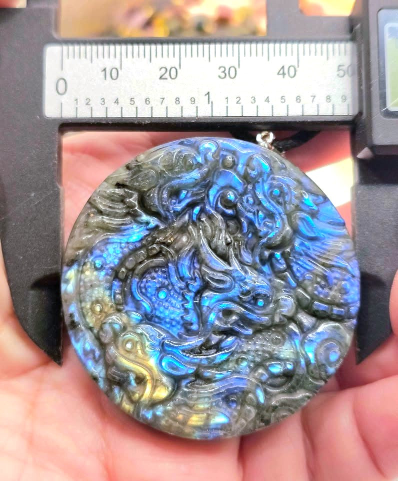 A close-up of a round carved stone pendant held in a hand under a caliper, showing a dragon design with strong blue, gray, and gold iridescent coloration.