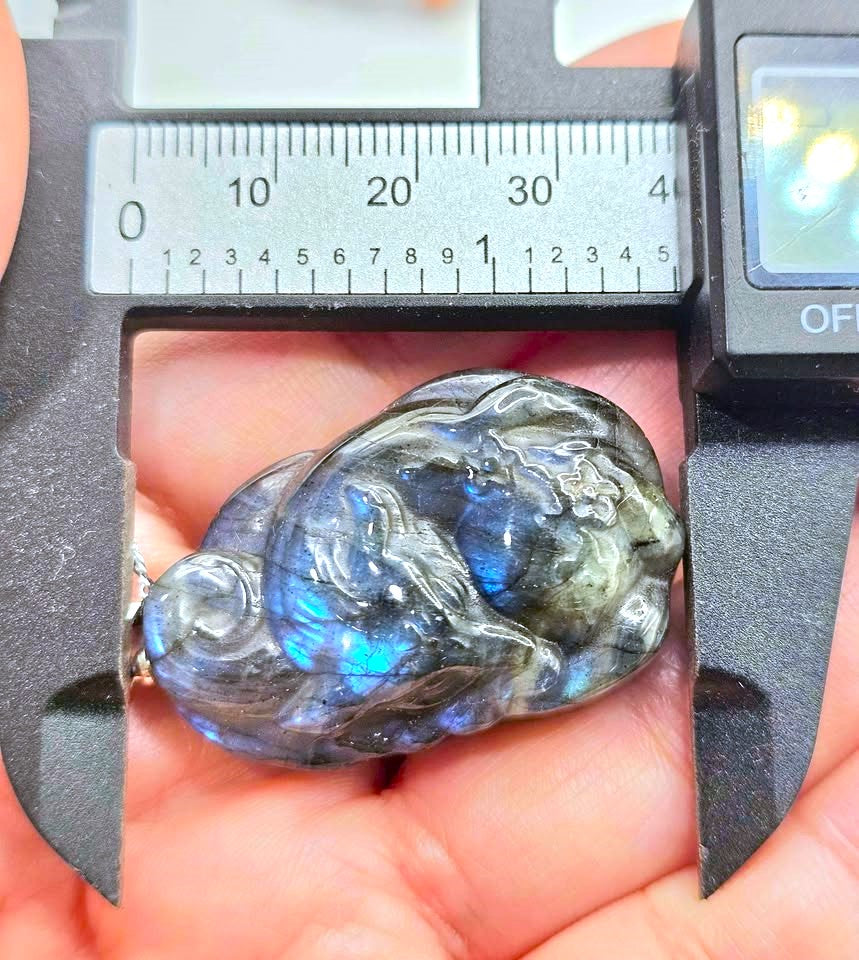 A polished labradorite pendant held in a hand under a caliper, showing its characteristic blue iridescent flash. The caliper is positioned to show the item's dimensions, and the background is the hand holding it.