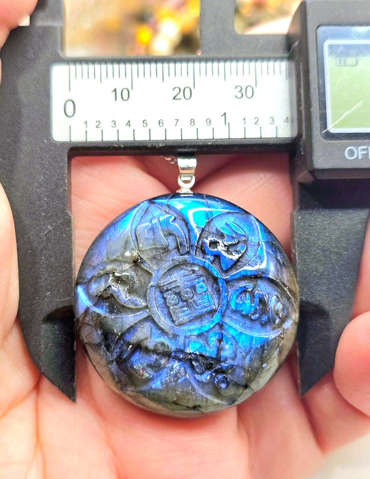 A round labradorite pendant with an engraved Chinese coin motif and blue iridescent flash, held in a hand under a digital caliper.