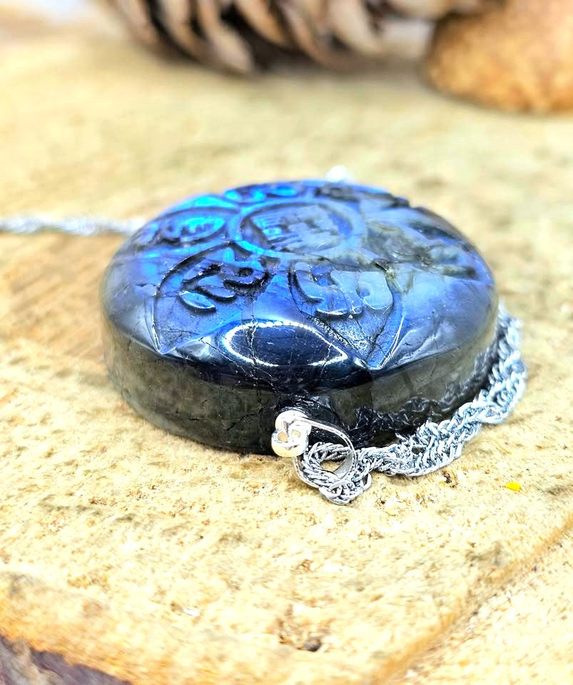 A round black carved pendant or locket with a silver chain, photographed close up on a textured tan surface with a blurred natural background.