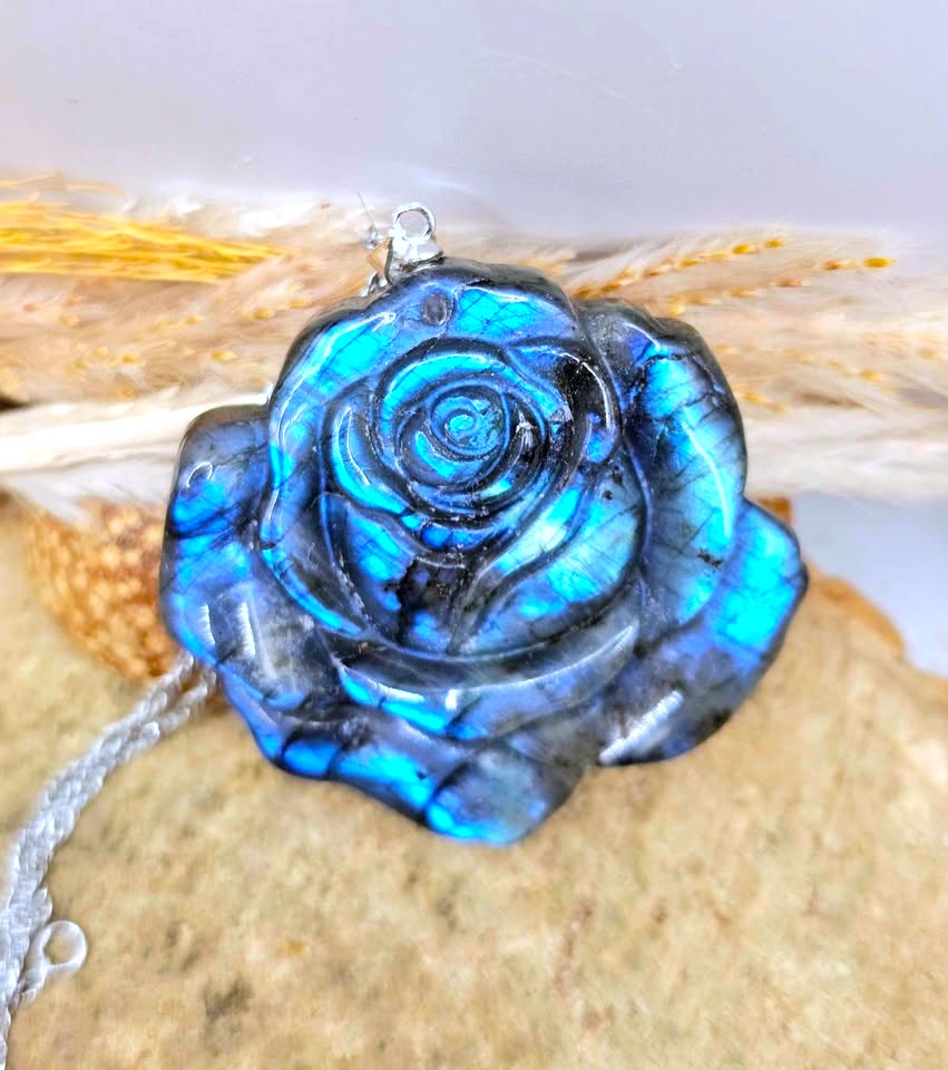 A blue labradorite rose-shaped pendant necklace with a silver bail, resting on a textured stone with dried wheat stalks in the background.