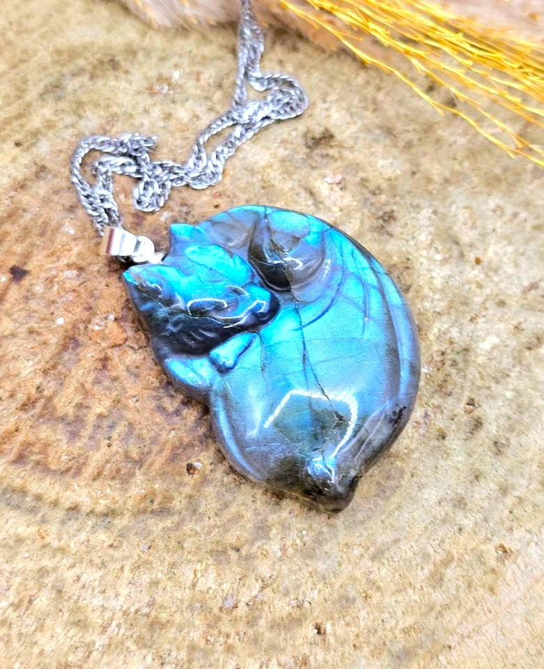 A polished cat-shaped pendant necklace with a silver chain, resting on a textured stone surface. The pendant features a vibrant blue-green iridescent finish with dark black and gray marbling.