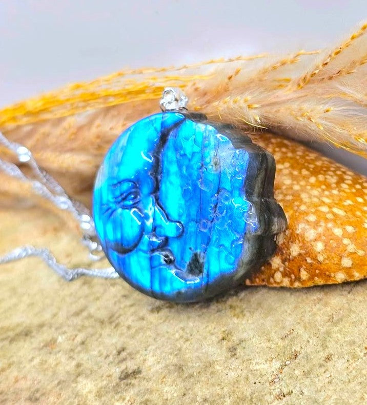 A round blue iridescent pendant necklace with a silver bail, resting on a textured sandy surface next to a dried orange feather.