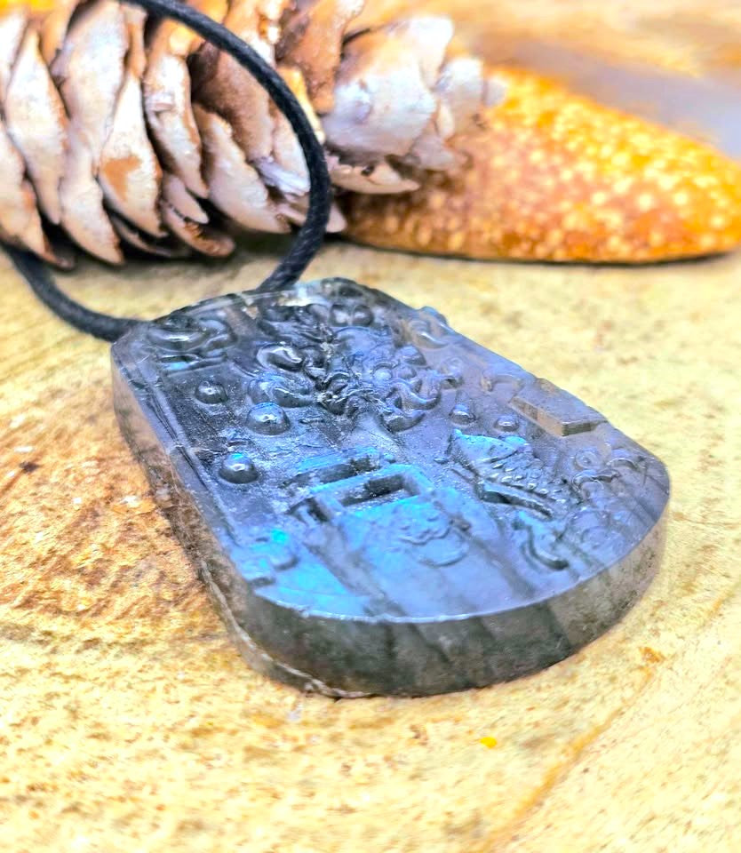 A dark gray carved stone pendant necklace with an embossed design, hanging from a black cord, displayed on a textured tan surface with a pinecone and starfish in the background.