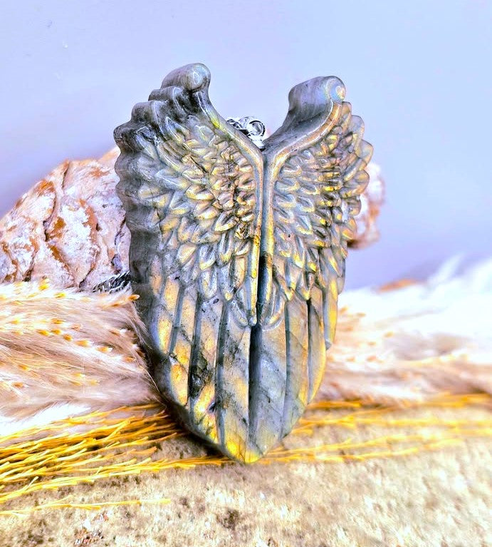 A carved winged shield pendant with textured feather-like detailing, shown standing upright on a decorative surface with dried grass and a small rock behind it.