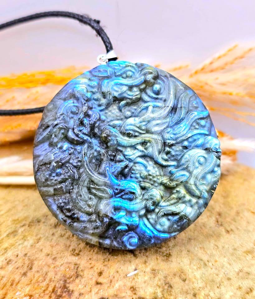 A round carved pendant necklace featuring a detailed dragon design with a strong blue iridescent sheen, suspended from a black cord, resting on a textured stone surface.