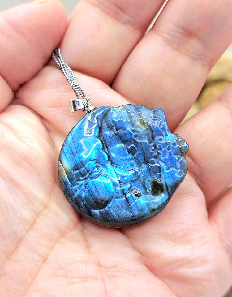 A round blue labradorite gemstone pendant with a silver chain, held in a person's hand to show its size and iridescent blue color play.