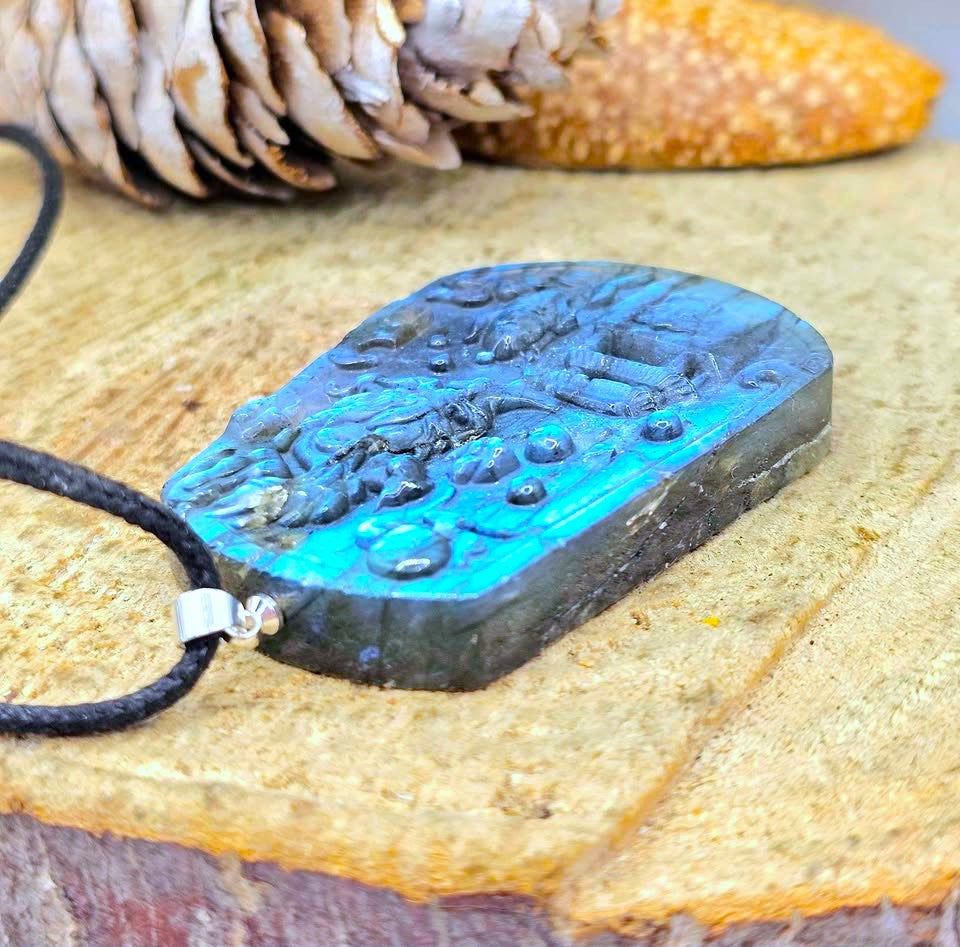 A carved rectangular pendant necklace with a black cord and silver clasp, featuring a blue-green iridescent stone surface and raised natural motifs, displayed on a textured wood surface with a pinecone and orange object in the background.