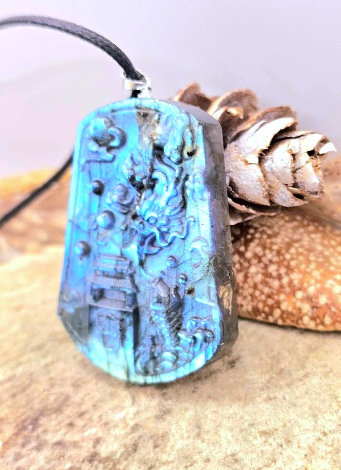 A rectangular blue flash carved pendant with a black cord, displayed on a sandy surface next to a pinecone.