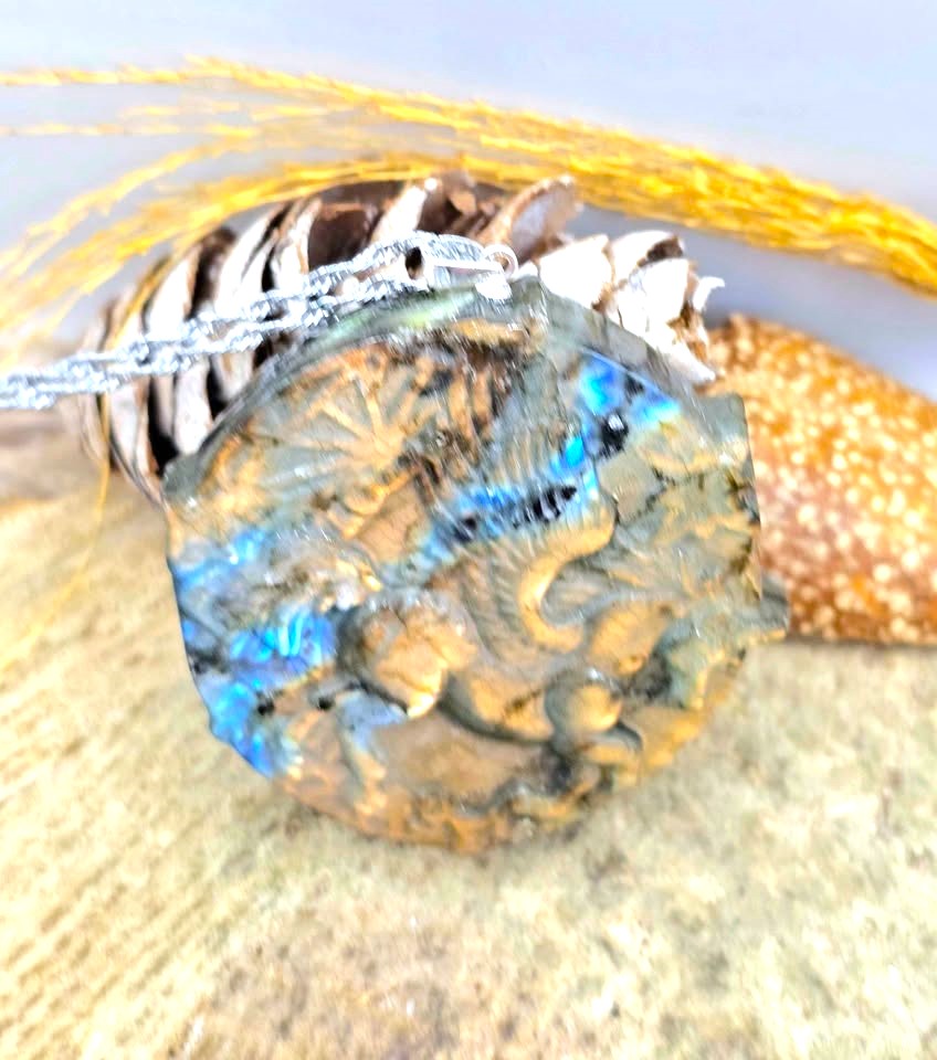A close-up of a round pendant necklace with a textured, metallic gold-toned surface and iridescent blue-green flashes, hanging from a silver chain over a decorative background of shells and gold thread.