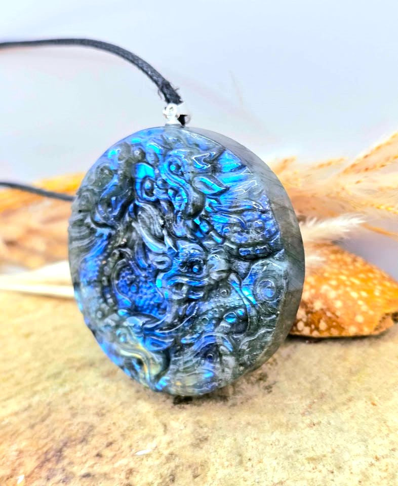 A round carved pendant necklace made of iridescent blue labradorite stone with a black cord, displayed on a sandy surface with dried grass and a shell in the background.