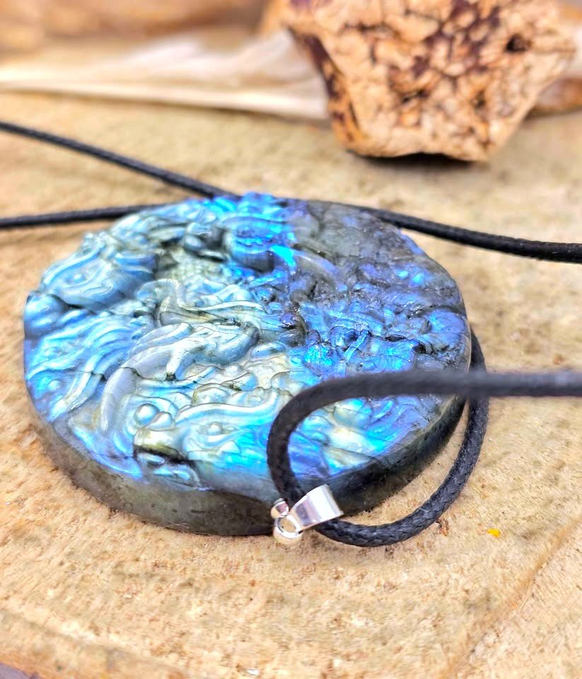A round carved pendant necklace with a black cord, featuring a blue and gray iridescent stone surface with an engraved figure, shown on a textured stone surface.