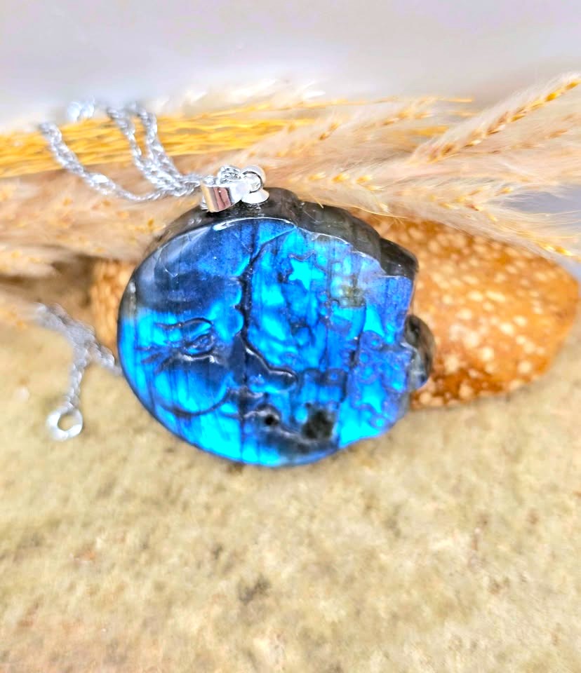 A round labradorite pendant necklace with a vibrant blue and black marbled pattern, hanging from a silver chain over dried wheat stalks on a sandy surface.
