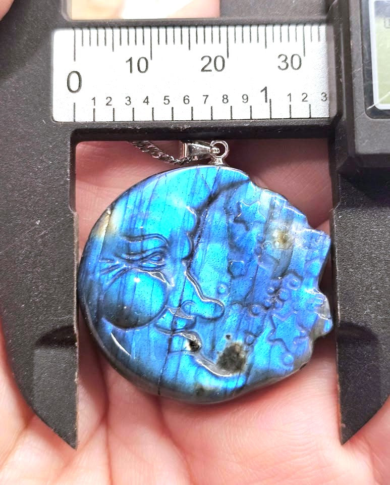 A round labradorite pendant with a blue iridescent sheen, held in a hand under a metal caliper with a ruler scale visible above it.