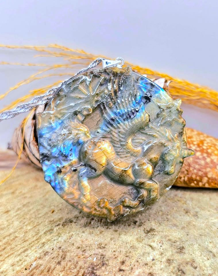 A round carved stone pendant featuring a detailed dragon motif, decorated with iridescent blue and gold labradorescence, resting on a textured stone surface with dried grass in the background.
