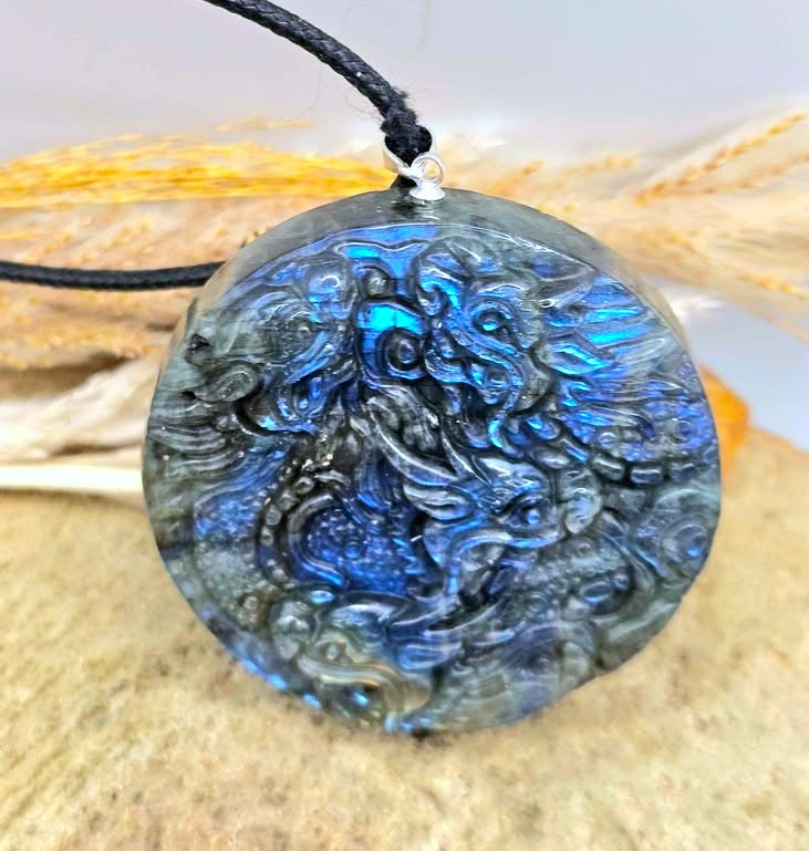 A round labradorite pendant necklace featuring a detailed carved dragon design with vibrant blue and green iridescent flashes, suspended from a black cord on a neutral background.