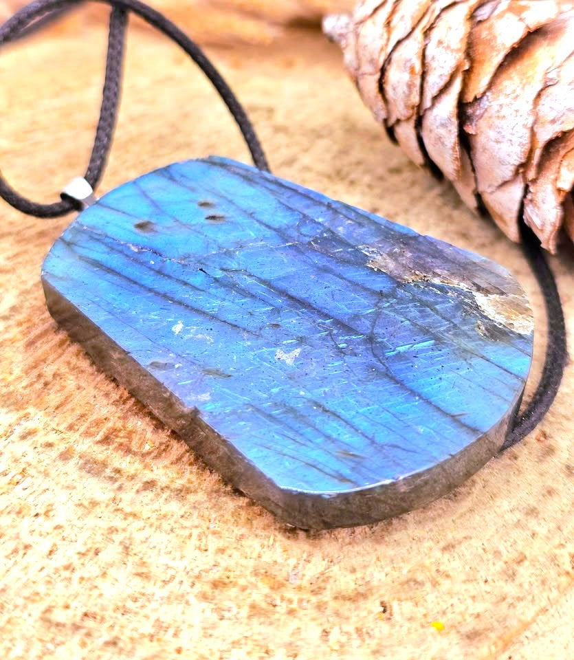 A raw labradorite stone pendant necklace with a black cord, displayed on a textured wood surface next to a pine cone.