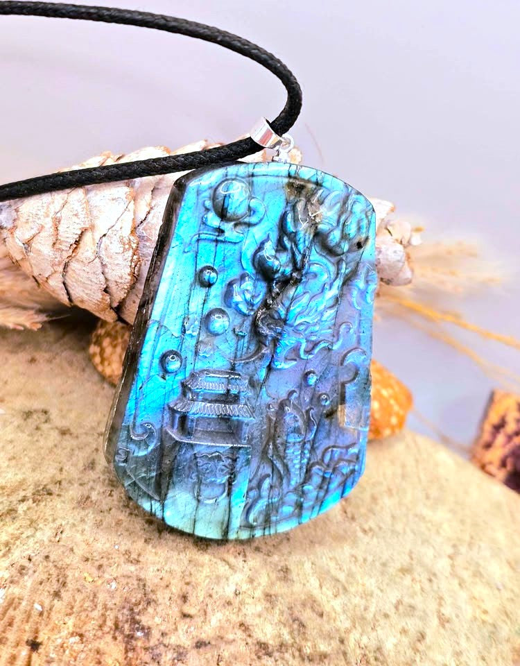 A rectangular pendant necklace with a black cord, featuring a highly textured, raised blue design resembling an dragon underwater scene. The pendant is displayed on a sandy surface next to a pine cone.
