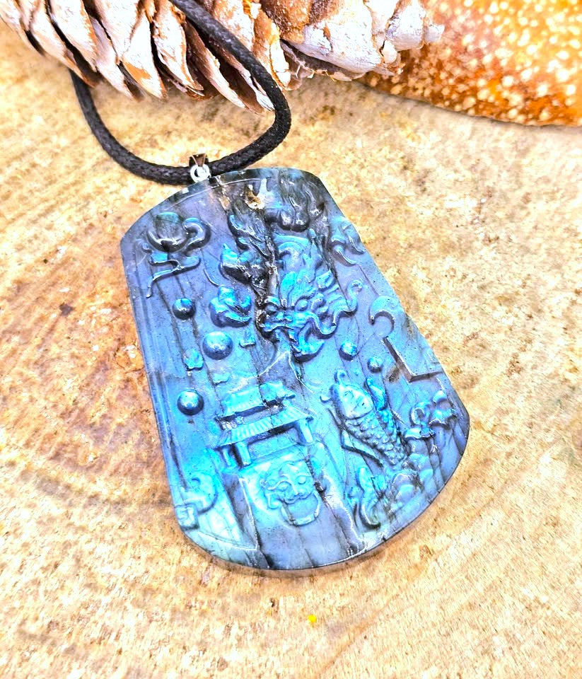 A carved blue-green stone pendant necklace with a black cord, displayed on a textured wood surface with a pinecone and orange decorative object in the background.