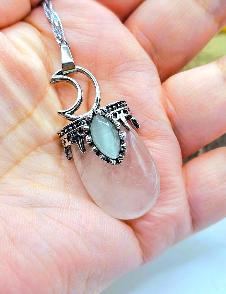 A close-up of a hand holding a silver pendant necklace with a crescent moon and ornate crown-like details surrounding a pale greenish-gray oval stone and a large teardrop-shaped translucent stone.