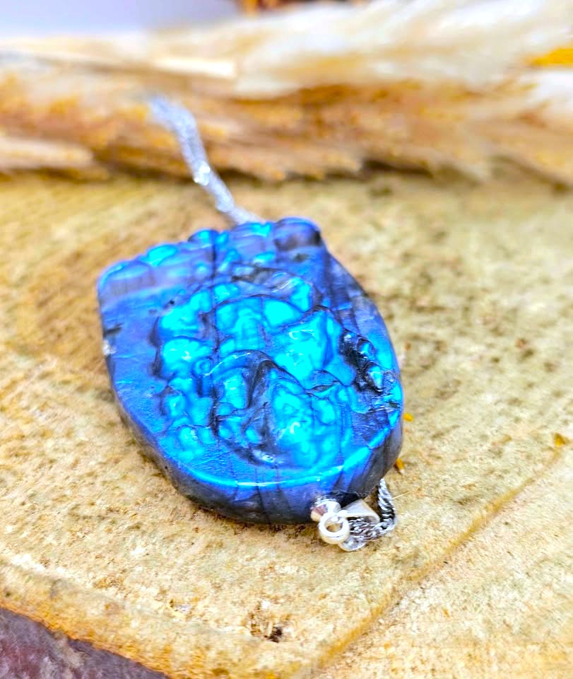 A close-up of a carved blue stone pendant with bright turquoise-like highlights and dark marbling, attached to a silver chain, resting on a textured tan surface.