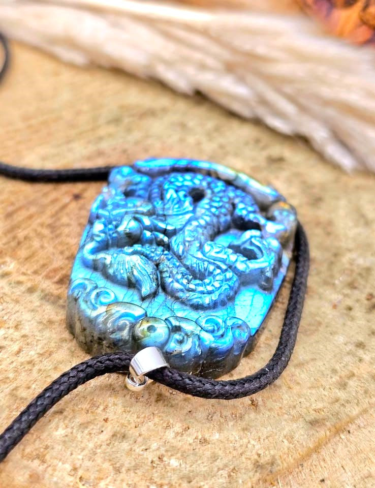 A carved blue dragon pendant necklace with a black braided cord and silver metal connector, displayed on a textured tan surface.