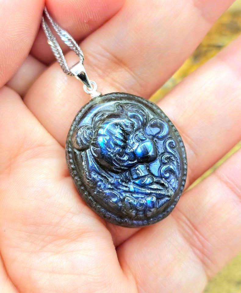 A close-up of a hand holding a silver pendant necklace with an oval dark stone featuring a raised classical face and blue iridescent highlights.