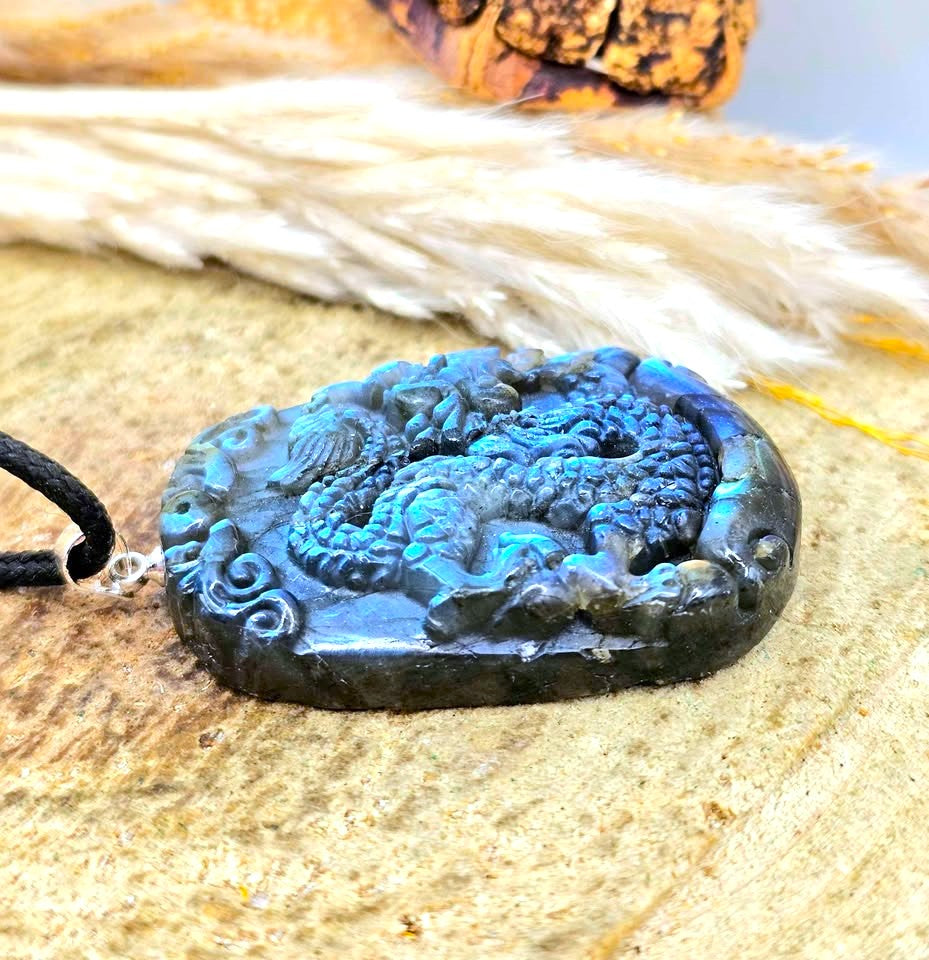 A carved stone pendant necklace featuring a detailed dragon design in vibrant blue and black, resting on a textured beige surface with dried grass in the background.