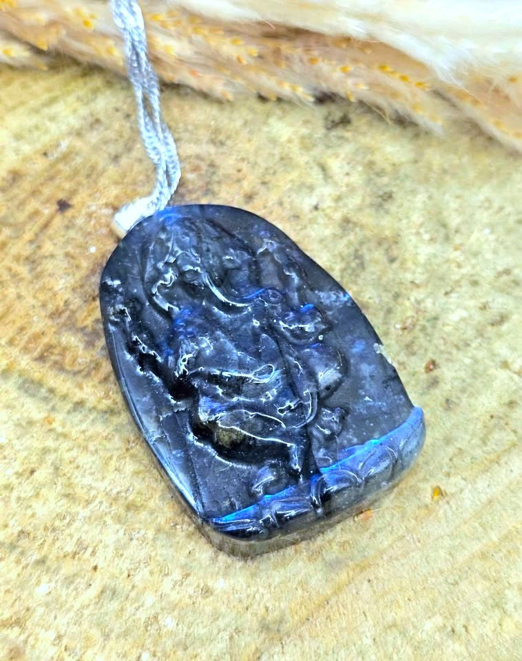A carved dark blue to black stone pendant with a light gray cord, photographed close-up on a textured tan surface.