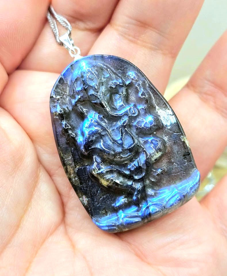 A hand holding a carved labradorite pendant with a silver bail. The pendant features a detailed relief carving of a figure, showcasing a strong blue flash and natural labradorite iridescence.