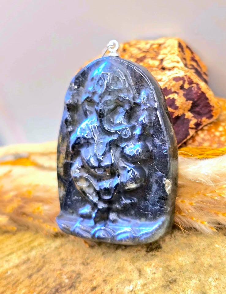 A carved black labradorite pendant featuring a detailed deity design, resting on a textured wooden surface with a gold-toned decorative object in the background.