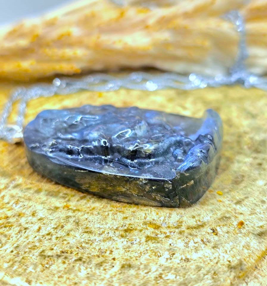 A raw, polished dark gray stone pendant with a rough, textured surface, resting on a textured beige stone surface with a silver chain partially visible in the background.