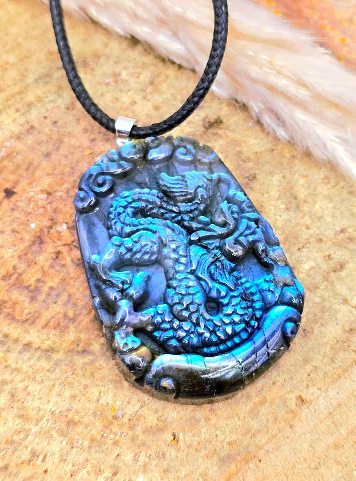 A carved dragon pendant necklace with a black cord, featuring a highly detailed dragon design in vibrant blue and black, resting on a textured tan surface.