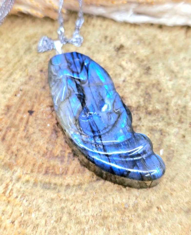 A polished labradorite pendant necklace with a silver-toned bail and chain, resting on a textured sandy surface. The pendant displays a vibrant blue and green iridescent play-of-color with dark natural matrix lines.