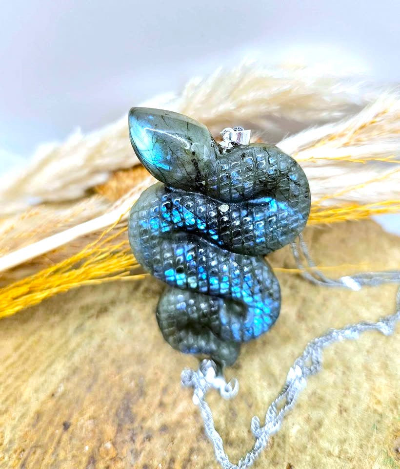 A coiled labradorite snake pendant with vibrant blue and green iridescent flashes, set in silver-tone metal and resting on dried grass over a textured tan surface.