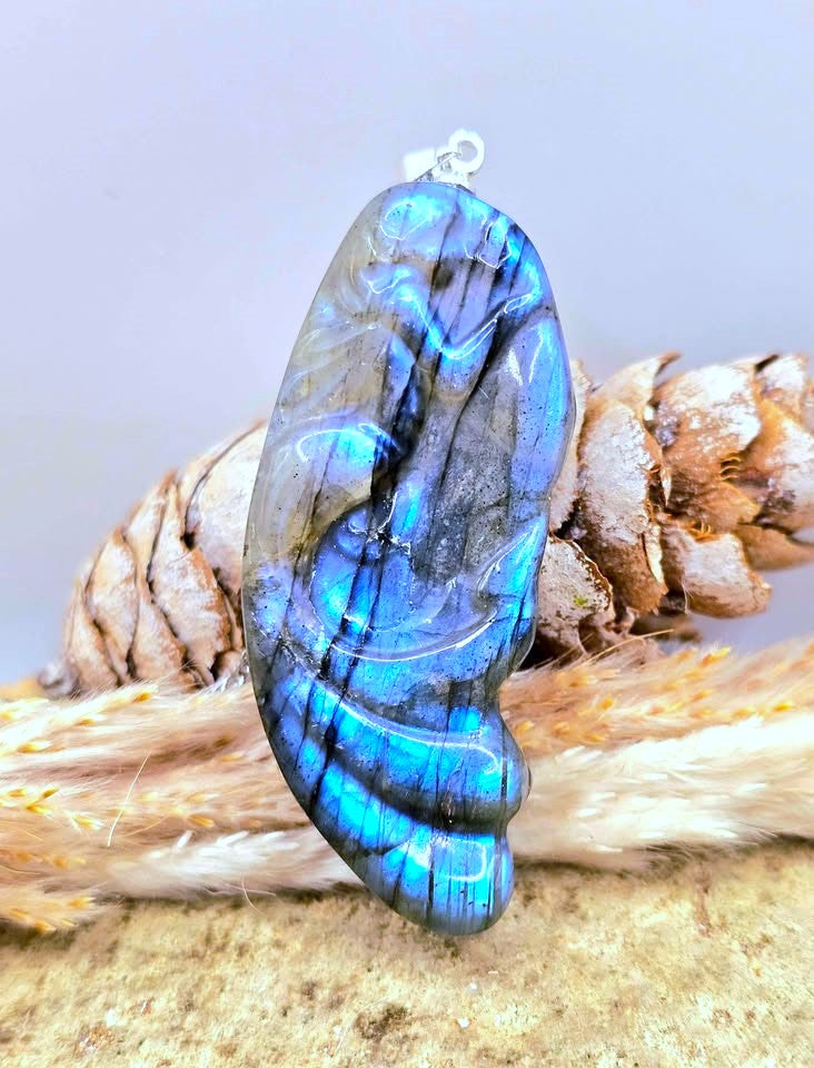A polished labradorite pendant with vibrant blue and green iridescent flashes, displayed on a bed of dried grass and pine cones against a soft gray background.