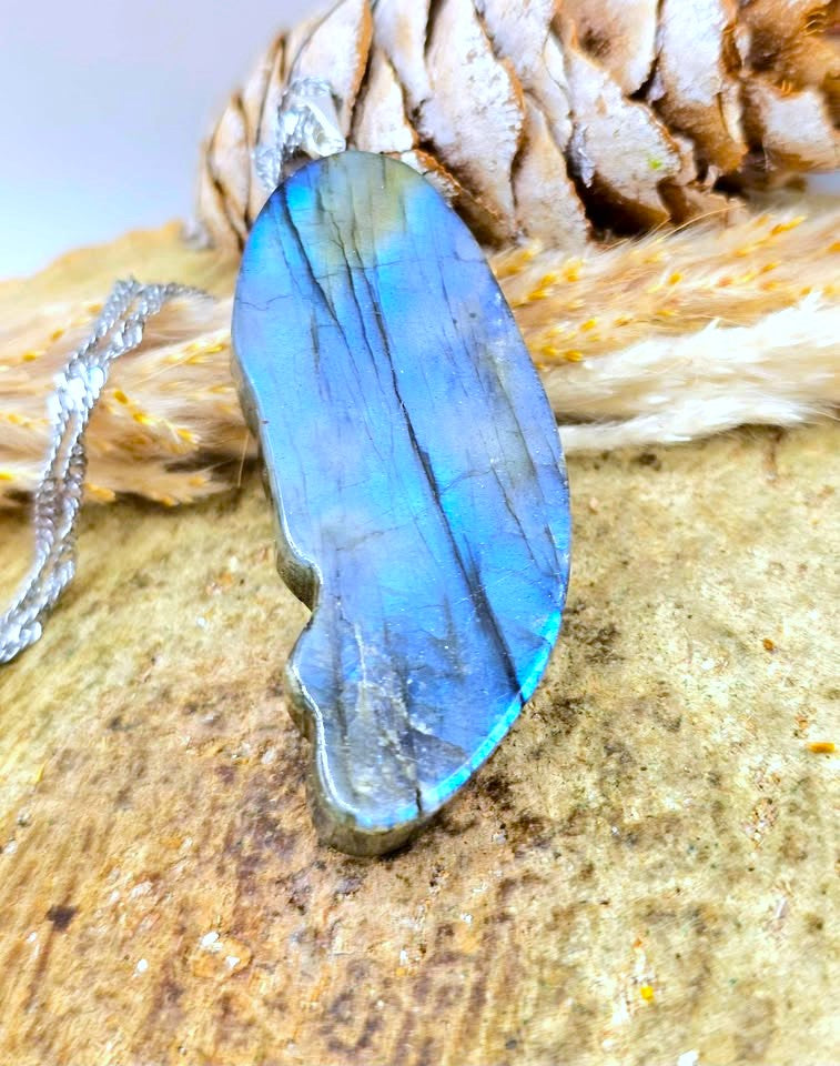 A polished labradorite pendant necklace with a silver-toned chain, resting on a textured wooden surface with a pinecone in the background.