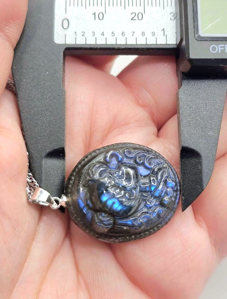 A close-up of a hand holding a round iridescent pendant with a silver chain, under a digital caliper to show its size. The pendant has a dark base with vibrant blue and green shimmering inclusions.