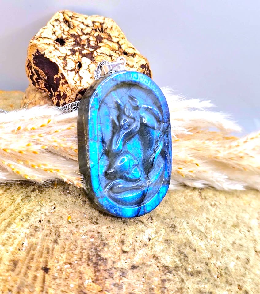 A carved oval pendant featuring a blue and black dragon design, displayed on a textured stone surface with dried grass and a decorative rock in the background.
