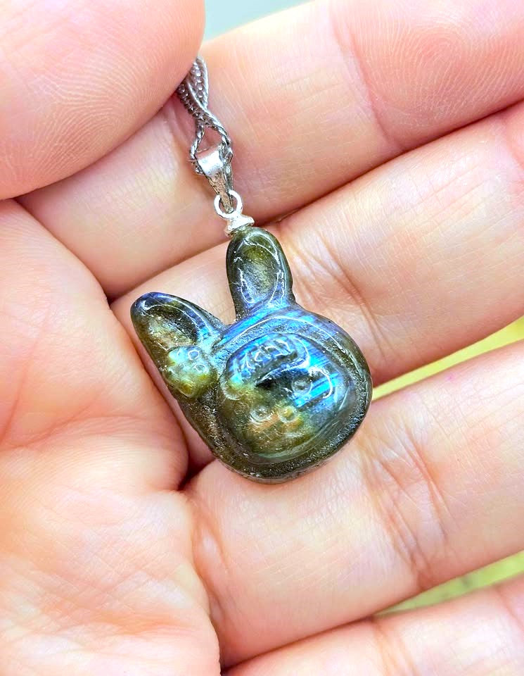 A close-up of a hand holding a silver bunny pendant with a highly iridescent, labradorescent green and blue finish.