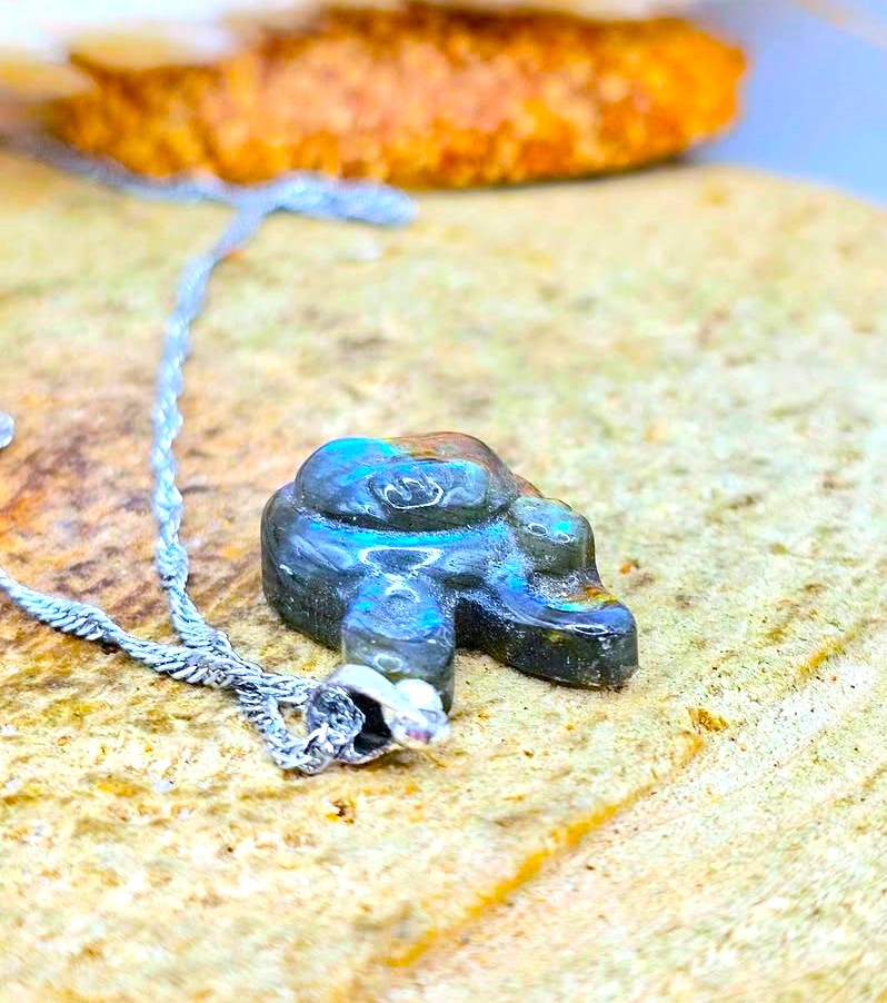 A small carved pendant or charm with a silver-toned chain, photographed close up on a textured stone surface. The pendant appears dark with iridescent blue-green highlights and a small clear stone attached near the bottom.