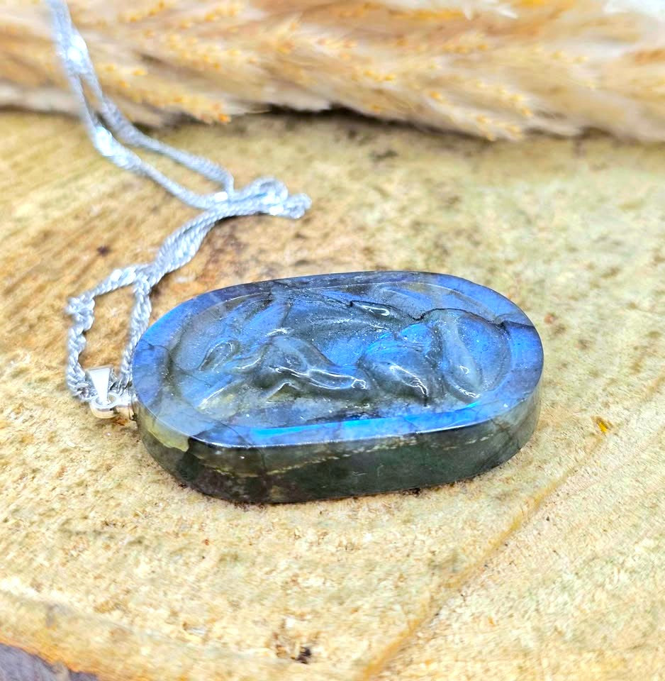 A carved oval labradorite pendant necklace with a silver-toned chain, resting on a textured stone surface.