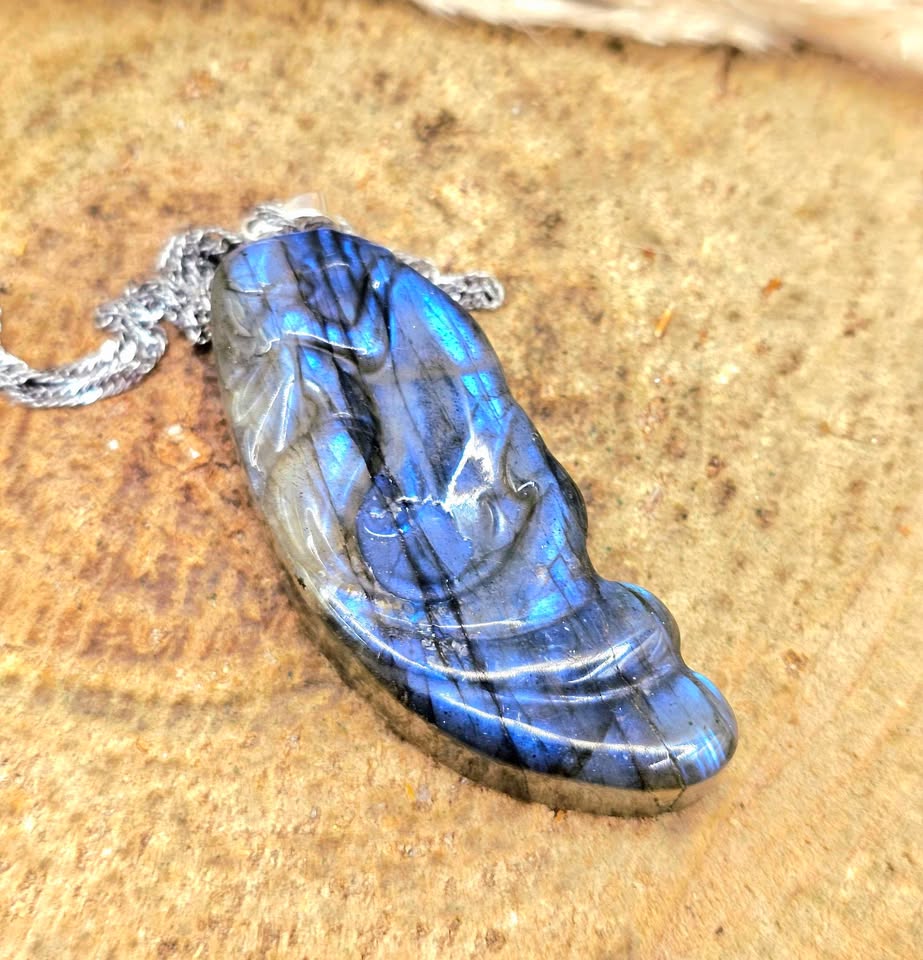 A polished labradorite pendant necklace with a silver chain, resting on a textured wood slice background. The pendant displays vibrant blue and gray flashes of light.