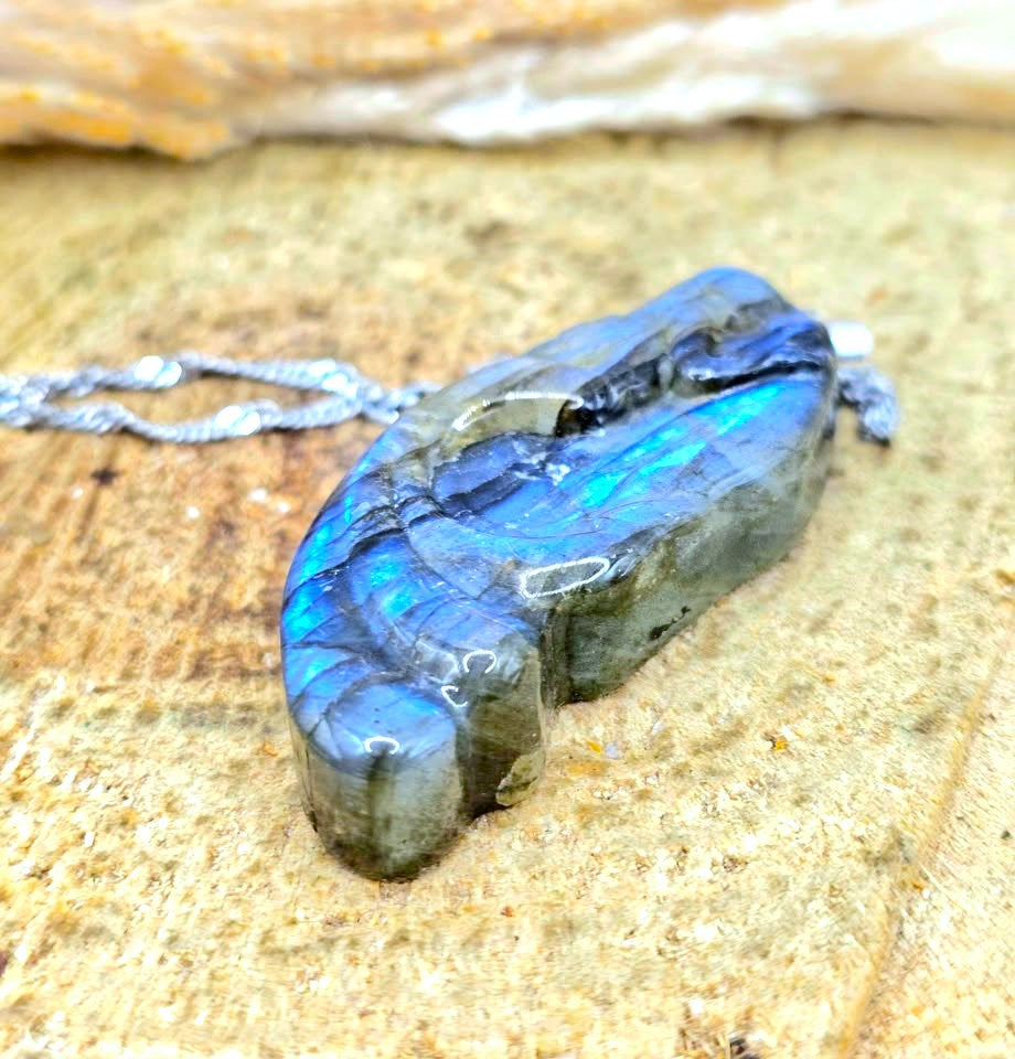 A polished labradorite pendant necklace with a silver chain, resting on a textured sandy surface. The labradorite displays vibrant blue and green flashes under natural light.