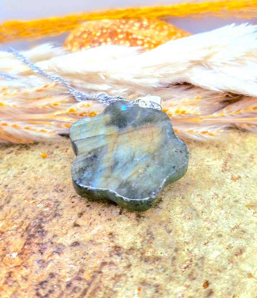 A polished labradorite stone pendant necklace with a silver chain, resting on a textured stone surface with dried grass and a shell in the background.
