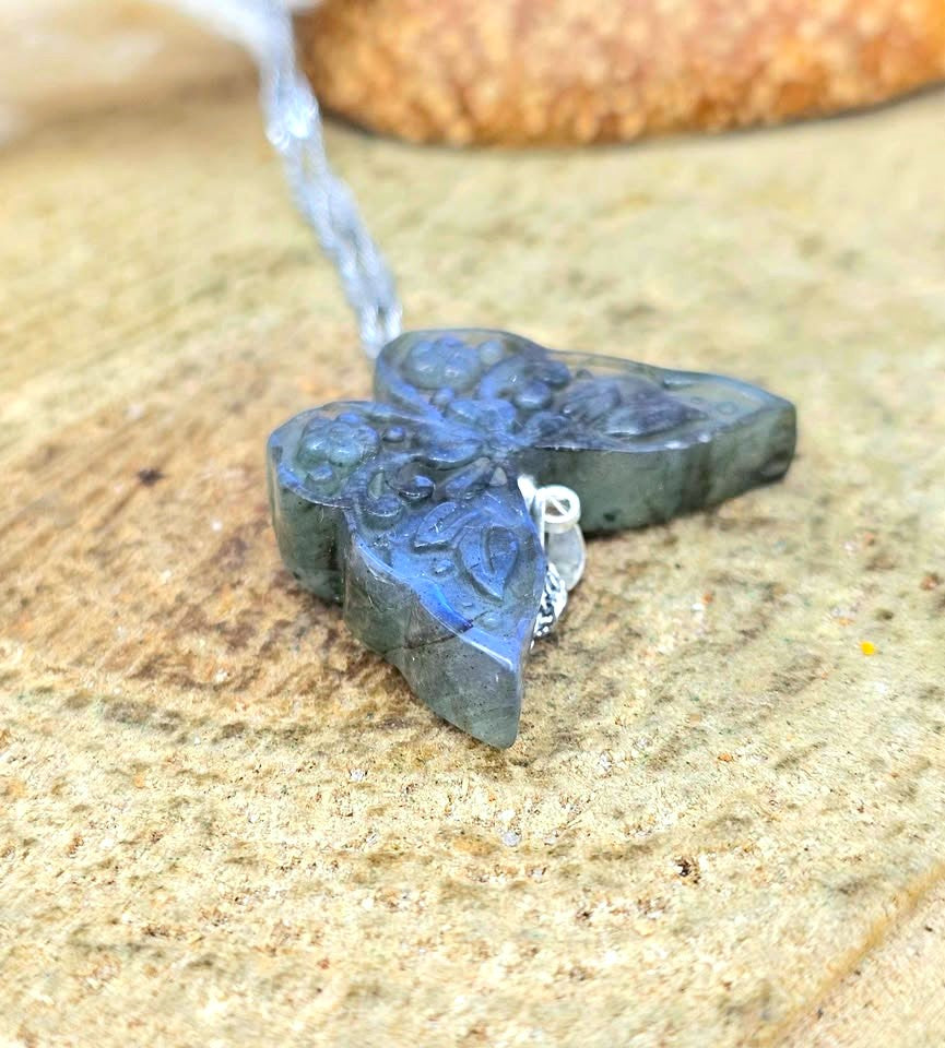 A close-up of a carved stone butterfly pendant in dark blue-gray with a silver-toned chain, resting on a textured sandy surface.