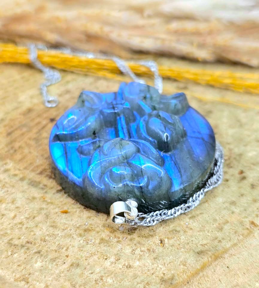 A carved labradorite pendant necklace with a silver chain, resting on a textured tan stone surface. The pendant displays vibrant blue and green iridescent labradorite flashes over a dark gray base.