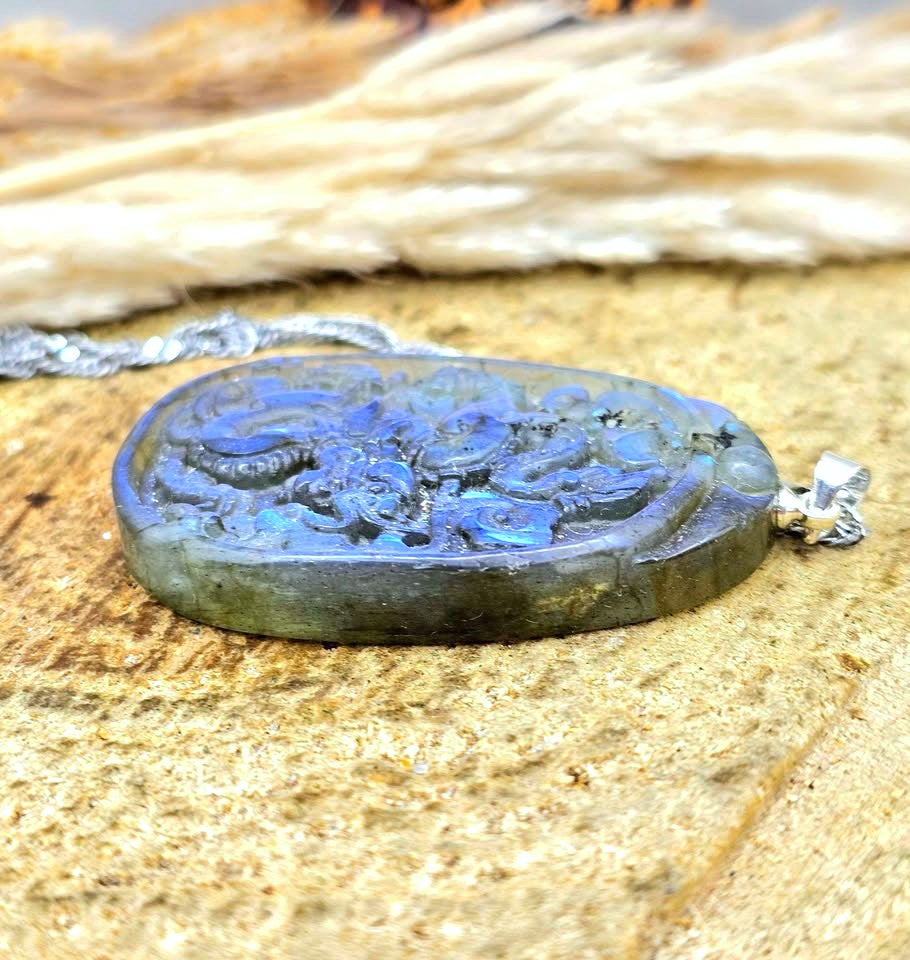 A carved oval labradorite pendant necklace with a silver bail, resting on a textured wooden surface with dried grass in the background.